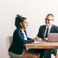 a female and a male sitting at a work desk smiling with laptop open. 